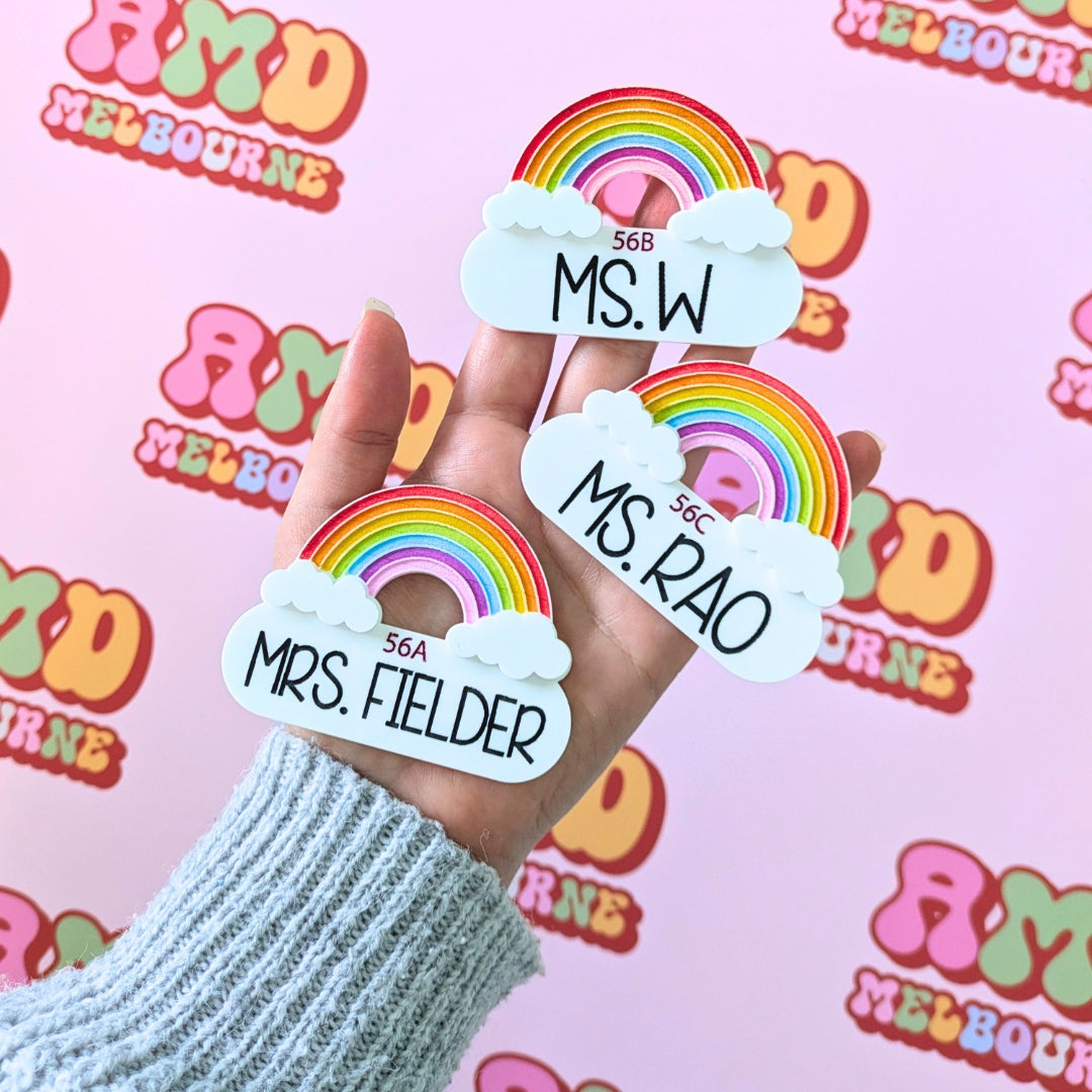 Hand holding 3 Acrylic Badges in the shape of rainbows with clouds against a pink AMDMelbourne monogram logo backdrop.